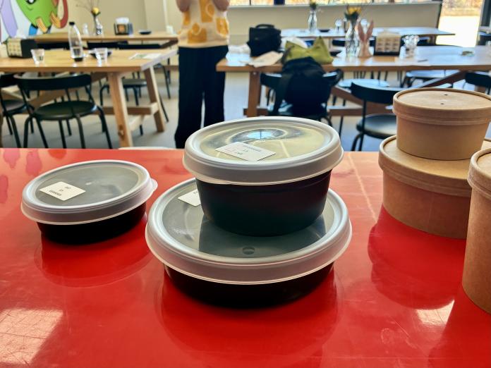 Nye bokse står stablet ovenpå hinanden på et rødt bord. I baggrunden står de gamle engangsbokse.