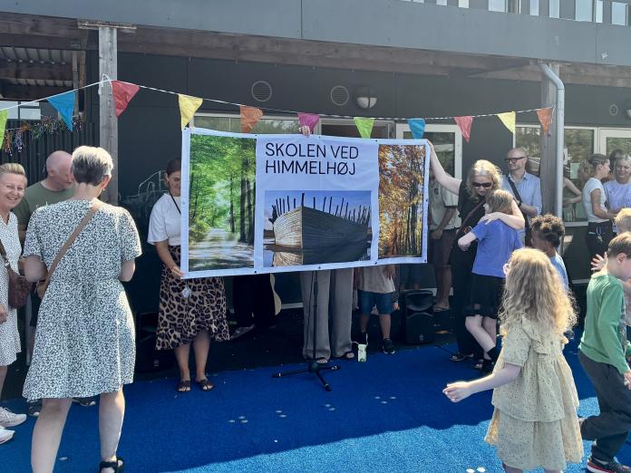 En gruppe mennesker foran et banner med teksten "Skolen ved Himmelbøj." Farverige flag hænger over dem.
